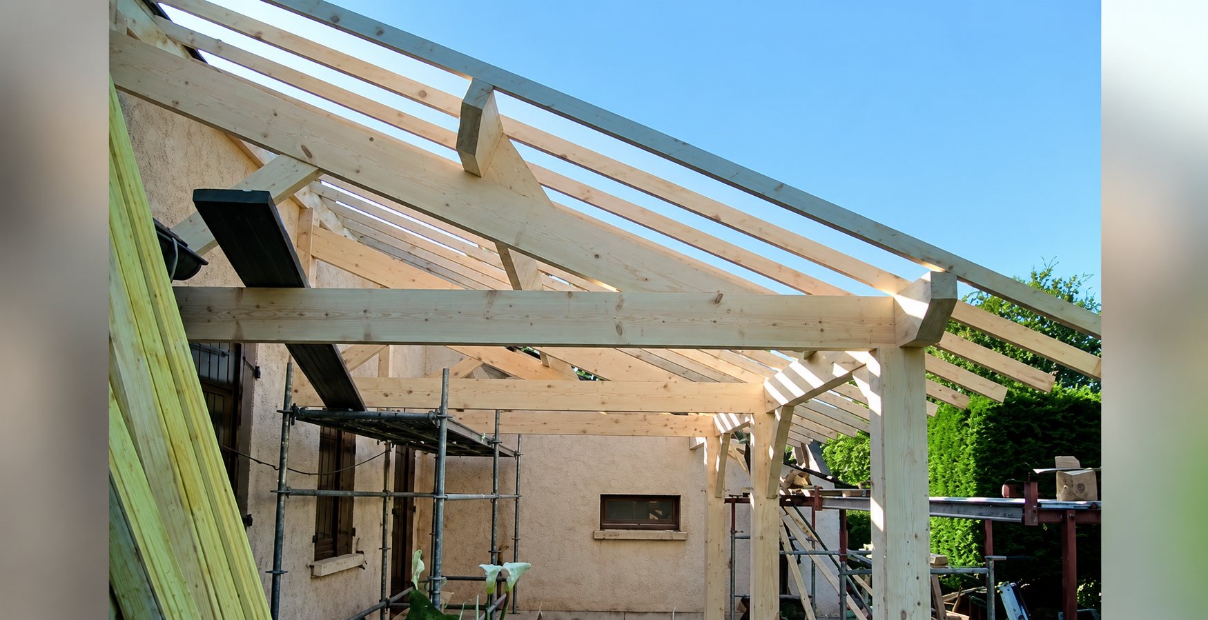 Structure de charpente en bois claire d’un auvent ou d’une toiture en cours de construction, adossée à une façade crépie, avec un échafaudage au fond.